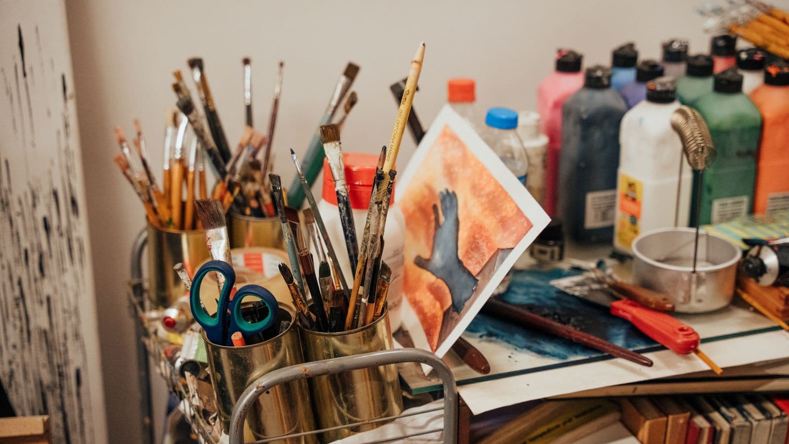 A crowded craft table with paintbrushes, paints, scissors and artwork